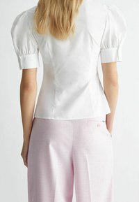 Weißes Oberteil mit Puffärmeln und strukturiertem Design. Glatte Stoffe, figurbetonte Taille und kurzärmelige Bündchen. Kombiniert mit hellrosa weitgeschnittenen Hosen.