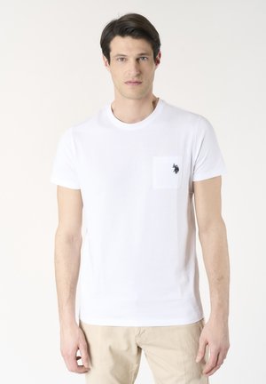 Giovane uomo con capelli corti e scuri che indossa una t-shirt bianca con un piccolo logo nero sulla tasca del petto e pantaloni beige.