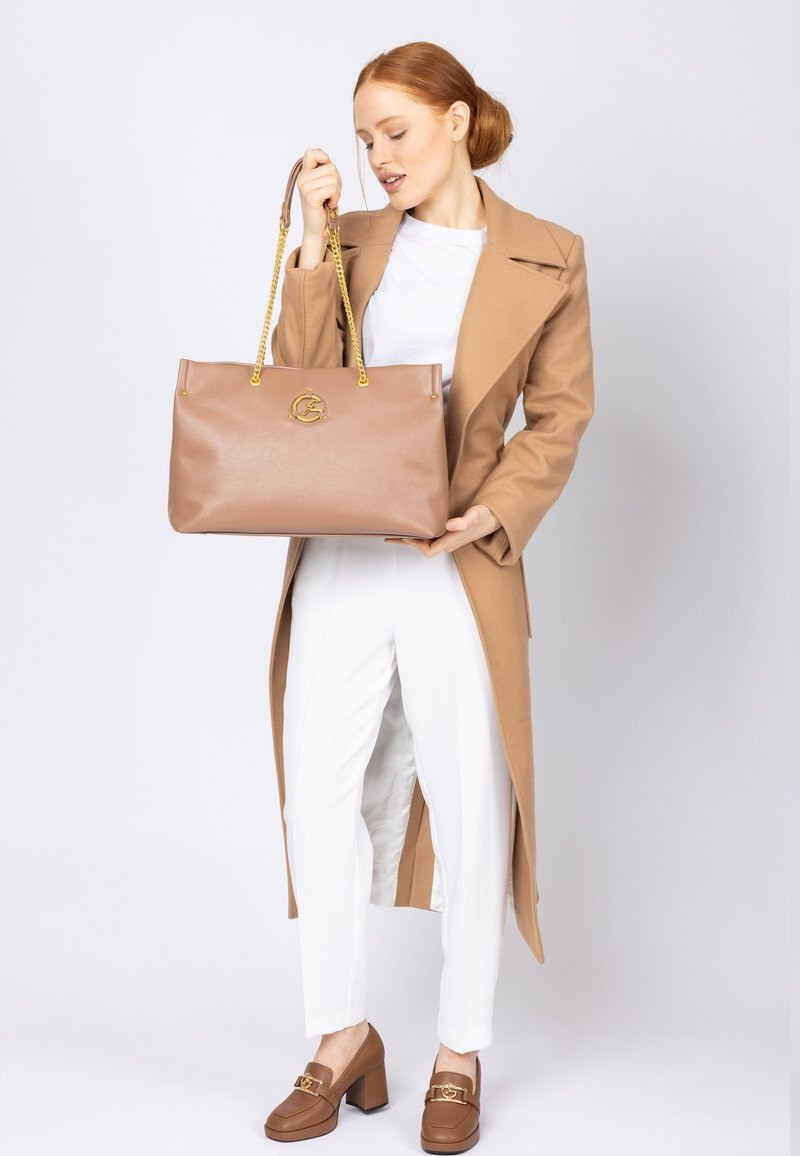 Bolso tote de piel marrón con acentos de hardware dorado y asas de cadena, que presenta un emblema central con el logo y un diseño suave y estructurado.