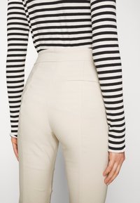 Béžové kalhoty na míru s hladkou texturou, vysokým pasem a zadní kapsou; v kombinaci s černobílým pruhovaným topem s dlouhým rukávem.