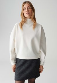 Weißer, übergroßer Pullover mit hohem Kragen aus weichem Material, kombiniert mit einem dunkelgrauen, anliegenden Rock. Glatte Textur und entspannte Silhouette.