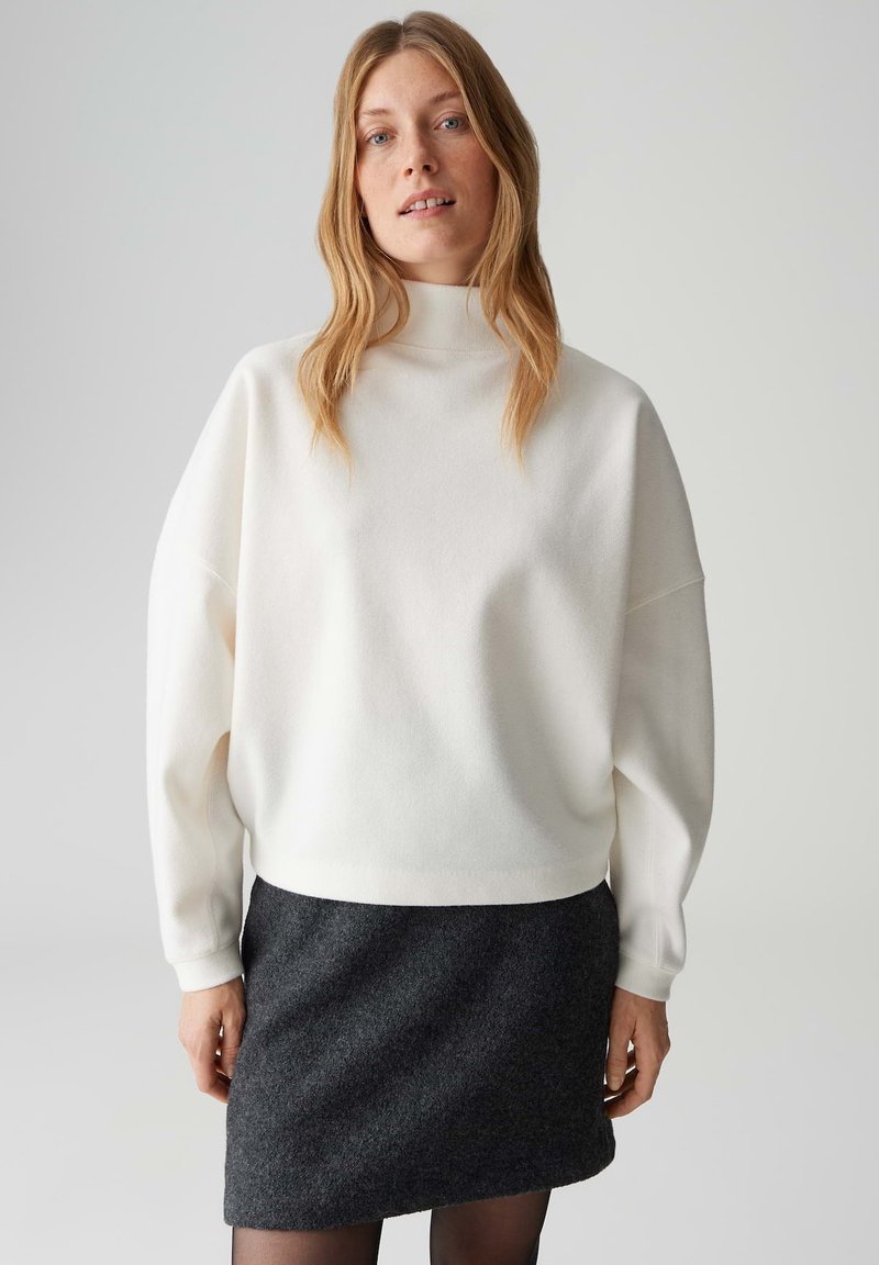 Weißer, übergroßer Pullover mit hohem Kragen aus weichem Material, kombiniert mit einem dunkelgrauen, anliegenden Rock. Glatte Textur und entspannte Silhouette.