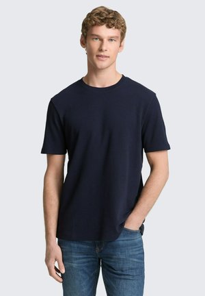 Młody mężczyzna o kręconych blond włosach, ubrany w granatową koszulkę z krótkim rękawem i niebieskie dżinsy, stoi z jedną ręką w kieszeni.