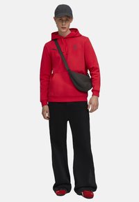 Rode hoodie met een logo, zwarte schoudertas, zwarte wijde broek en rode schoenen. Een grijze pet maakt de outfit compleet.