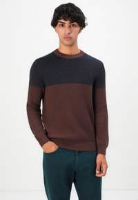 Jeune homme aux cheveux bouclés et foncés, portant un pull côtelé bi-couleur bleu marine et marron, avec une main dans la poche, se tenant contre un fond blanc.