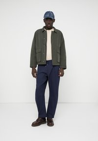 Man wearing green jacket, beige shirt, navy pants, brown shoes, and blue patchwork cap standing against white background.