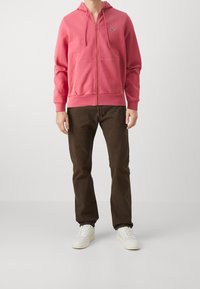 Felpa con cappuccio rosa con zip, tasca a marsupio e cappuccio con coulisse, abbinata a pantaloni marroni e sneakers bianche. Tessuto liscio e design casual.