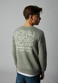 Giovane con felpa verde adornata da un design floreale astratto bianco e la scritta "Sinfonia delle Emozioni" sulla schiena, in posizione di profilo.