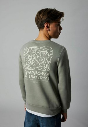 Jeune personne portant un sweat-shirt vert avec un motif floral abstrait blanc et le texte "Symphony of Emotions" dans le dos, se tenant de manière latérale.