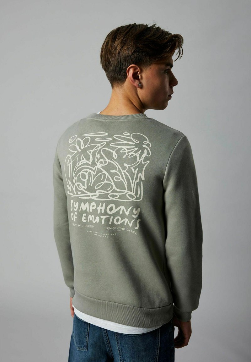 Giovane con felpa verde adornata da un design floreale astratto bianco e la scritta "Sinfonia delle Emozioni" sulla schiena, in posizione di profilo.