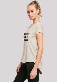 Beige cotton t-shirt with a round neckline and short rolled sleeves, featuring black text "Manifest the Patriarchy" and a relaxed fit.
