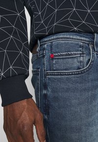 Black sweatshirt with white geometric pattern, paired with blue denim jeans. Features a small red accent on the back pocket.