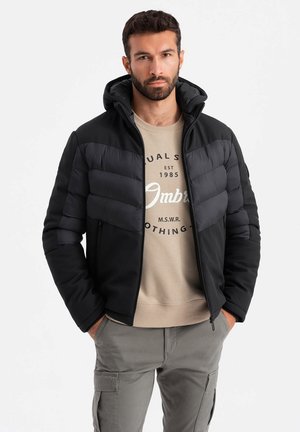 Sort pufferjakke med hætte, der har en glat stofoverflade, quiltede sektioner og sidelommer med lynlås, lagd ovenpå en beige sweatshirt.
