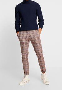 Pull col roulé côtelé navy, pantalon à carreaux marron avec des accents jaunes et blancs, associé à des baskets montantes beiges.