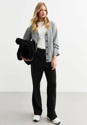 Grijze cardigan met knopen, witte t-shirt, zwarte broek en witte sneakers. Houdt een zwarte, gestructureerde deken vast. Casual outfit.