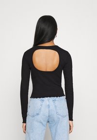 Schwarzes Langarmoberteil mit großem Ausschnitt auf der Rückseite. Strukturierter Stoff, körpernaher Schnitt, gewellter Saum, kombiniert mit hellblauen Jeans.