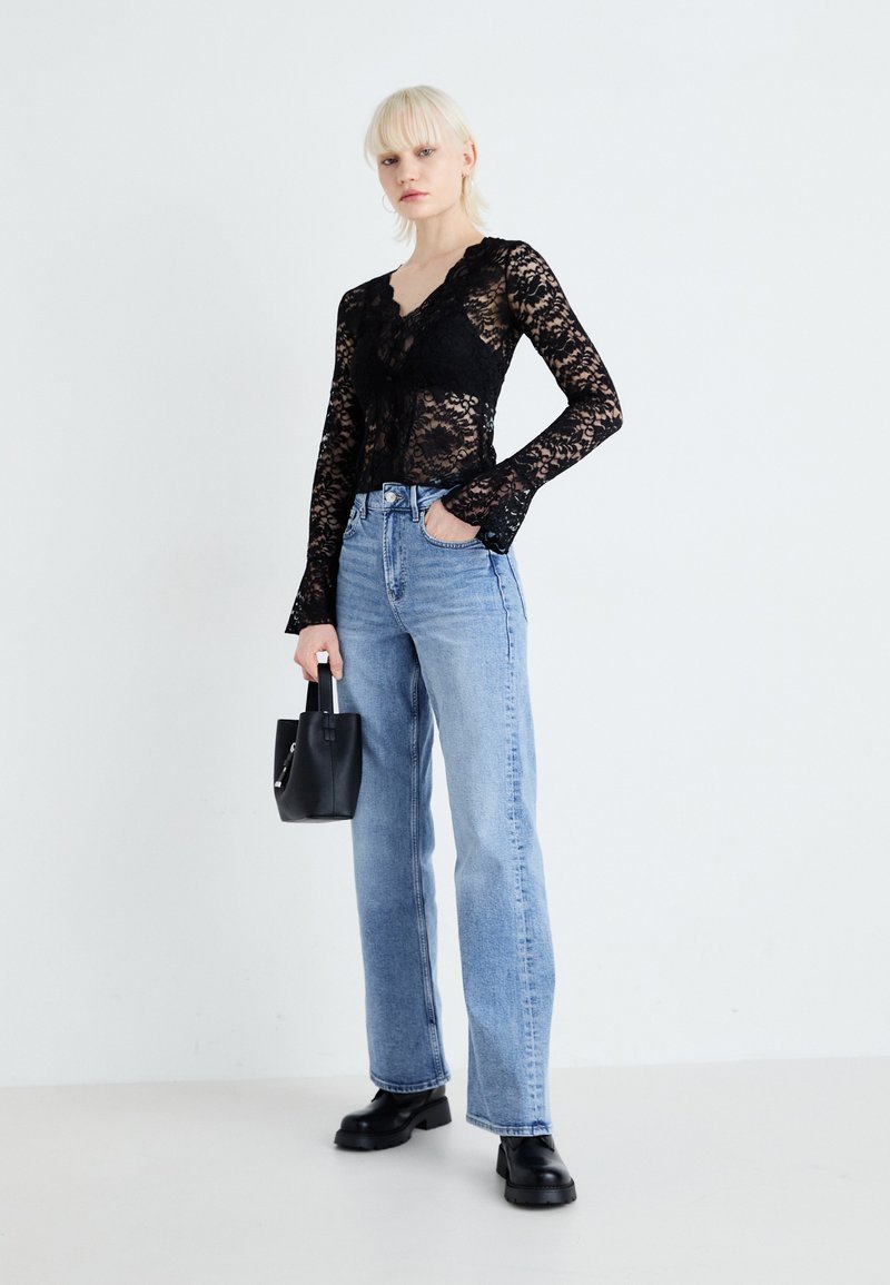 Haut en dentelle noire à manches longues avec un col festonné, associé à un jean droit bleu clair. Le mannequin tient un petit sac à main noir et porte des bottes noires épaisses.