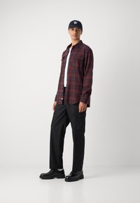 Koszula w kratę flanelowa w kolorze czerwonym i granatowym, czarne spodnie cargo i czarne buty. Model nosi granatową czapkę z białym logo. Prosty design, luźny fason.