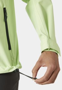 Chaqueta verde clara con un bolsillo con cremallera, que cuenta con un dobladillo ajustable con cordón y un tejido suave y elegante. Mano tirando del cordón.