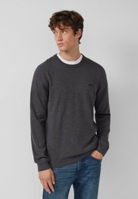 Giovane uomo con capelli ricci castani che indossa un maglione grigio scuro sopra una camicia bianca e jeans blu, che guarda verso sinistra con una mano in tasca.
