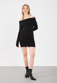 Vestido negro de punto acanalado, con tirantes caídos, de mangas largas, diseño ajustado y longitud hasta el muslo, combinado con botas de tacón puntiagudas negras.
