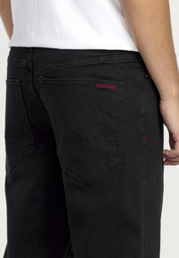 Jeans noirs avec une texture lisse, comportant des coutures rouges et une étiquette de marque sur la poche arrière. Le design inclut des poches standards et des coutures.