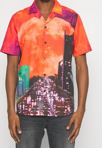 Chemise à manches courtes avec boutons, présentant un imprimé galaxie vibrant en rouge et orange, une silhouette de ville noire, et des bâtiments verts contrastants. Tissu lisse.