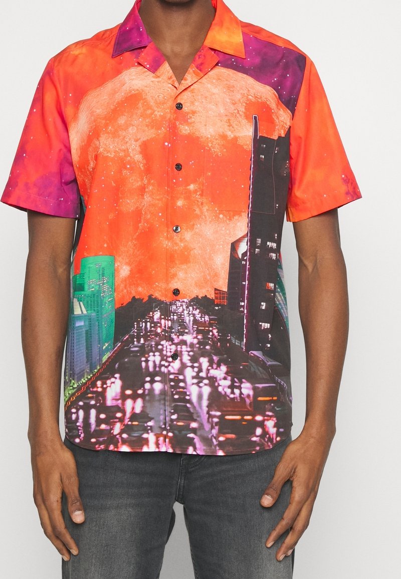 Chemise à manches courtes avec boutons, présentant un imprimé galaxie vibrant en rouge et orange, une silhouette de ville noire, et des bâtiments verts contrastants. Tissu lisse.