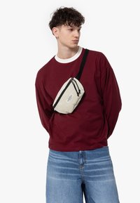 Borsa a tracolla a righe beige con zip nera, indossata sopra una maglietta a maniche lunghe color bordeaux. La borsa è dotata di una tracolla e di un dettaglio con logo.