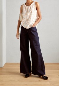 Haut blanc à fleurs avec un col volanté, associé à un jean large en denim foncé. La personne porte des chaussures à plateforme noires et brillantes.