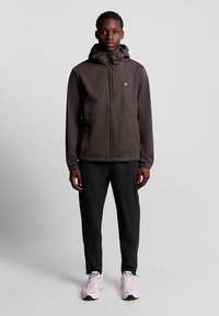 Donkergrijze hoodie met een frontale rits, elastische manchetten en een klein gele logo. Gecombineerd met zwarte broeken en lichte sneakers.