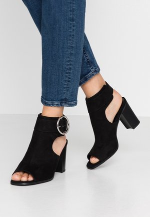 Zapatos de tacón abiertos de ante negro con un tacón grueso y un acento de hebilla circular de plata en el tobillo. Altura de tacón ligera y diseño inclinado.