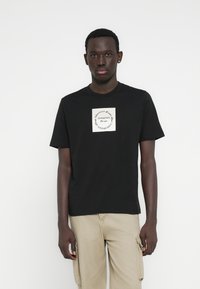 Μαύρο βαμβακερό μπλουζάκι με στρογγυλή λαιμόκοψη και κοντά μανίκια, που διαθέτει κρεμέ λογότυπο στο στήθος. Απλός σχεδιασμός και εφαρμογή.