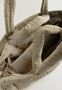 Un sac fourre-tout beige texturé avec des détails cousus, montrant le tissu intérieur et une poche zippée, mettant en avant son design spacieux.