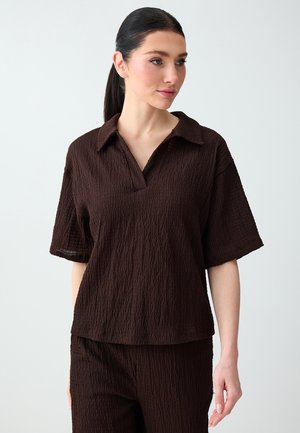 Vrouw met donker haar in een lage paardenstaart, gekleed in een donkerbruine, textuurrijke korte mouw blouse met kraag en bijpassende broek, kijkt opzij.