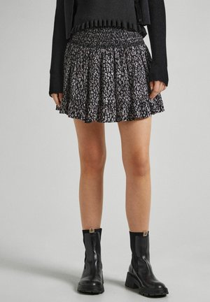 Personne portant une mini-jupe noire à motifs, un haut noir à manches longues et des bottines noires, debout devant un fond neutre.
