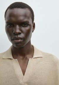 Beige texturovaný pletený svetr s výstřihem do V a žebrovaným límcem, nošený osobou s tmavou pletí a krátkými vlasy, na pozadí s jednobarevným pozadím.