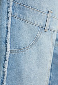 Vaaleansininen farkkukangas, jossa on purkautunut reuna. Siinä on sivutasku, vyölenkit ja sävyyn sopivaa ommelta yksityiskohtana. Rakenteellinen, rento muotoilu.