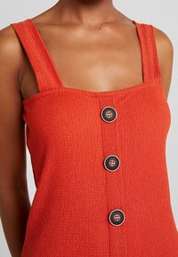 Punainen teksturoitu maksimekko, jossa on paksut olkaimet, neliönmuotoinen pääntie ja kolme tummaa nappia edessä yksityiskohtana.