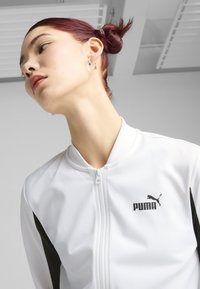 Bijela jakna s patentnim zatvaračem i crnim detaljima, napravljena od glatkog materijala. Sadrži Puma logo na lijevoj strani prsnog koša i dizajn sa uspravnim ovratnikom.