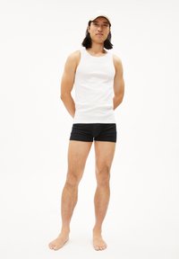 Witte katoenen tanktop en zwarte shorts op een mannelijk model dat blootvoets staat, met armen gekruist achterin. Eenvoudig ontwerp en effen kleuren.