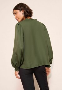 Sötétzöld blúz áttetsző anyagból, puffos ujjal és ráncolt nyakkivágással, karcsú szárú fekete nadrággal párosítva. Sima textúra.