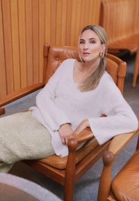 Jasnoszary prążkowany sweter i cekinowa beżowa spódnica, siedzi na drewnianym krześle z skórzanym siedziskiem, na tle drewnianej boazerii.