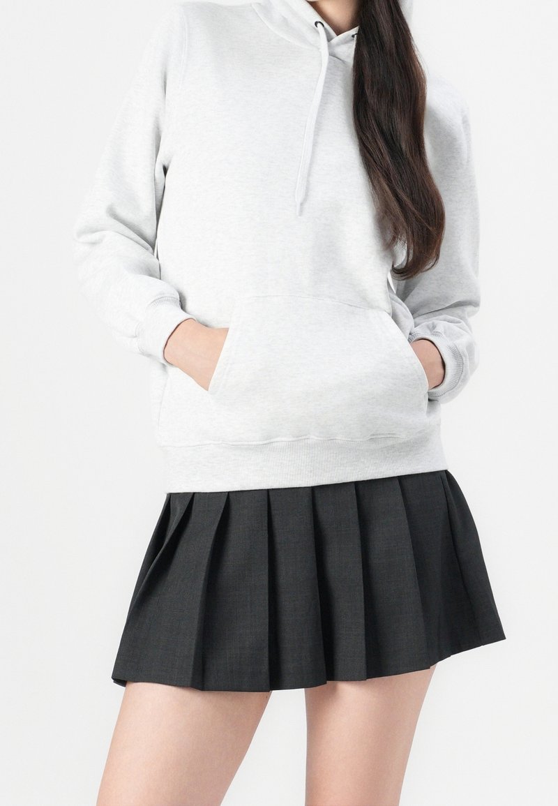 Personne portant un sweat à capuche gris clair, les mains dans la poche avant, et une jupe plissée courte noire sur un fond uni.