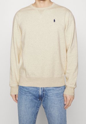 Uomo che indossa una felpa beige a maniche lunghe con un piccolo logo scuro sul petto, abbinata a jeans blu.