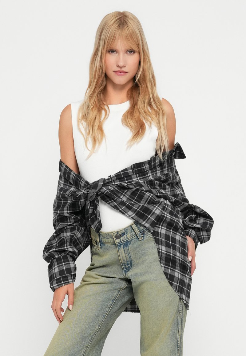 Camicia a quadri nera e grigia con bottoni legata in vita sopra una canotta bianca, abbinata a jeans verde sbiadito.