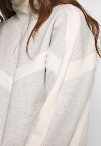 Un sweat-shirt gris clair avec un col montant, une silhouette structurée, et des bandes crème contrastantes sur les manches pour plus de détail et de texture.