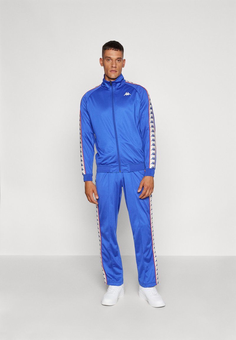Blauwe trainingspak van glad, lichtgewicht materiaal. Voorzien van een ritsjack met gestreepte zijkanten en bijpassende broek, geaccentueerd met een logo.