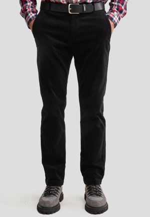 Pantalons en velours côtelé noir à coupe slim, avec poches latérales et une texture lisse, associés à des chaussures en daim grises et une ceinture noire.