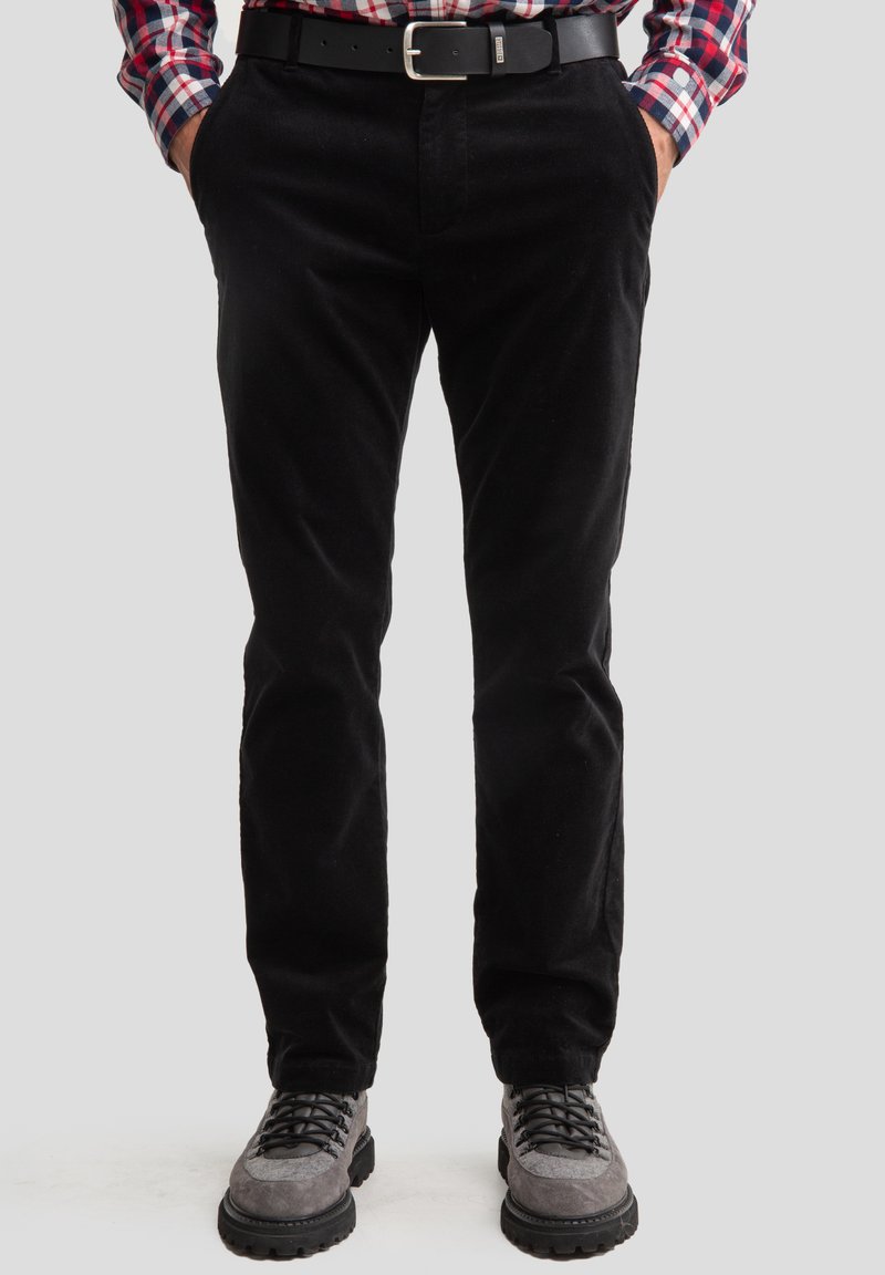 Pantalons en velours côtelé noir à coupe slim, avec poches latérales et une texture lisse, associés à des chaussures en daim grises et une ceinture noire.